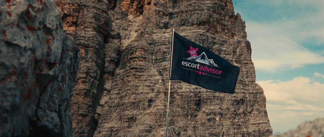 Bandiera di Escort Advisor che sventola davanti a una delle Tre Cime di Lavaredo, sotto un cielo azzurro con nuvole